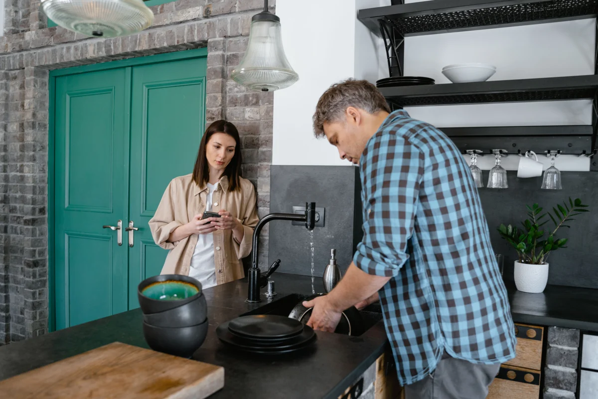 A man washing dishes beside another person in a lived-in kitchen